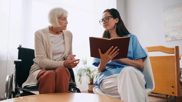 Брюнетка-медсестра в очках читает книгу счастливой пожилой женщине в инвалидном кресле — стоковое фото