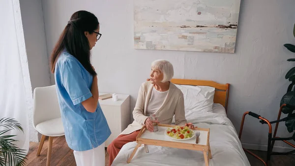 Enfermera morena hablando con paciente anciano cerca de bandeja con desayuno en la cama - foto de stock