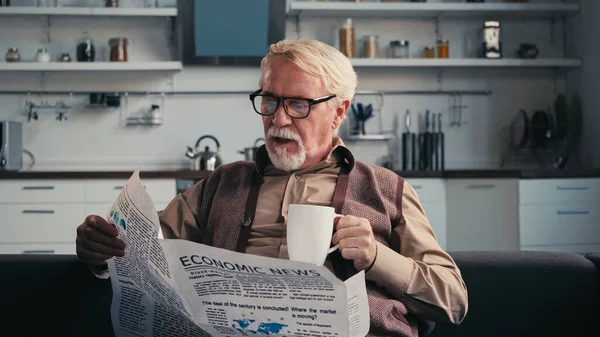 Homme âgé dans les lunettes de lecture journal et tenant une tasse de thé à la maison — Photo de stock