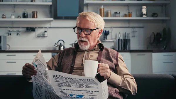 Senior man in eyeglasses reading newspaper and holding cup of tea at home — Stock Photo