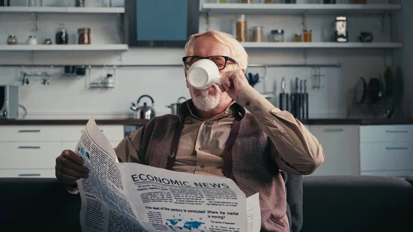 Senior mit Brille hält Zeitung in der Hand, während er zu Hause Tee trinkt — Stockfoto