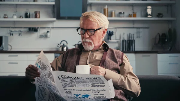Uomo anziano in occhiali che legge giornale mentre tiene la tazza di tè — Foto stock