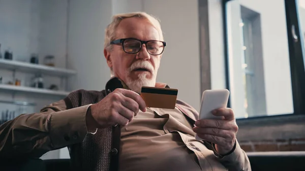 Старший мужчина с кредитной картой держит мобильный телефон во время покупок в Интернете дома — стоковое фото
