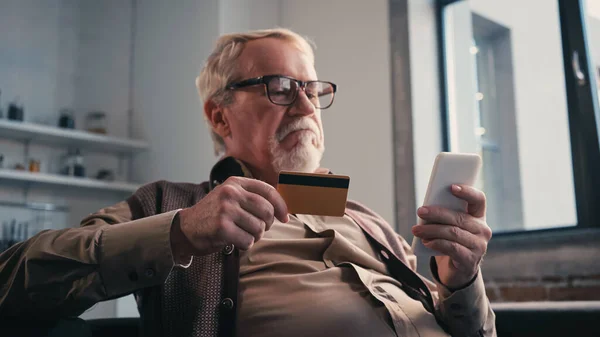 Hombre mayor disgustado con tarjeta de crédito celebración de teléfono celular mientras que las compras en línea - foto de stock