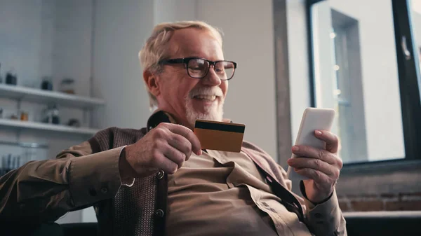Homme âgé gai avec carte de crédit tenant téléphone portable tout en faisant des achats en ligne à la maison — Photo de stock