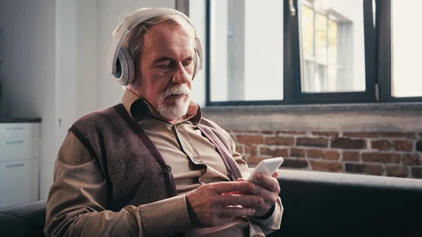 Hombre barbudo y mayor en auriculares inalámbricos escuchando música y utilizando el teléfono inteligente en casa — Stock Photo