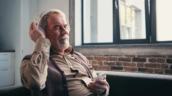 Hombre satisfecho y mayor en auriculares inalámbricos escuchando música y sosteniendo el teléfono inteligente en casa - foto de stock