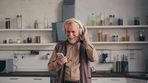 Счастливый пожилой человек в беспроводных наушниках, слушая музыку и держа смартфон на кухне — стоковое фото