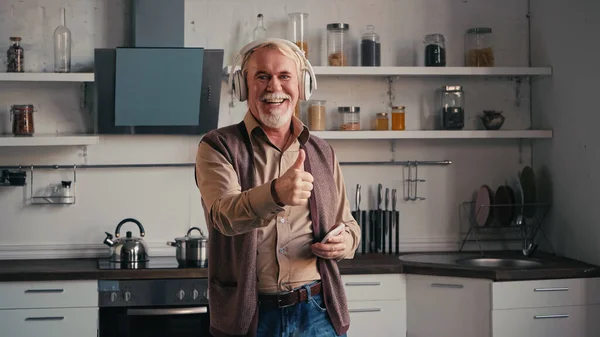 Happy and senior man in wireless headphones listening music, holding smartphone and showing thumb up in kitchen — Stock Photo