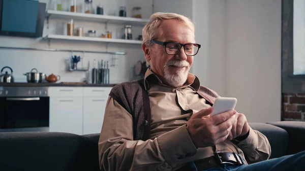 Homem idoso alegre em óculos mensagens no celular em casa — Fotografia de Stock