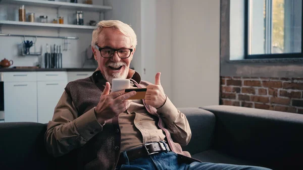 Happy pensioner with credit card holding mobile phone while showing thumb up at home — Stock Photo