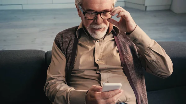 Retired man adjusting eyeglasses and looking at smartphone — Stock Photo