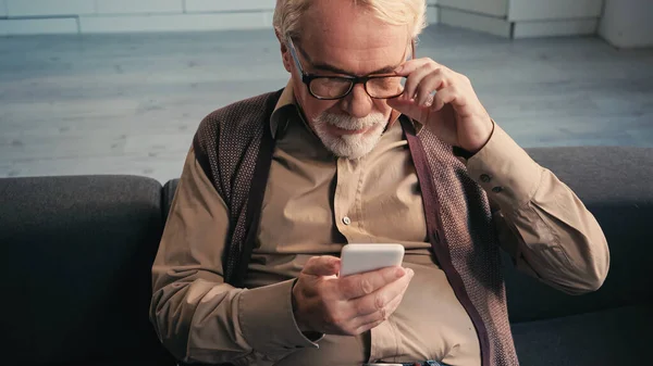 Bearded pensioner adjusting eyeglasses and looking at smartphone — Stock Photo