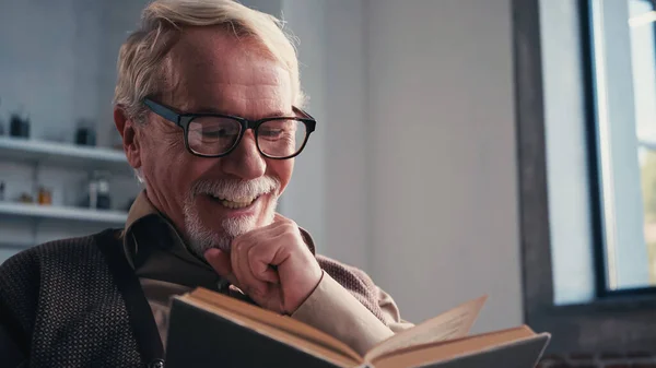 Homem idoso alegre em óculos lendo livro em casa — Fotografia de Stock