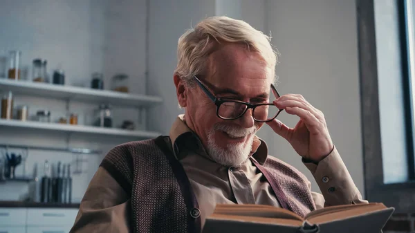 Positive senior man adjusting eyeglasses and reading book at home — Stock Photo