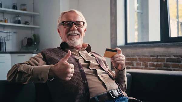 Щасливий пенсіонер в окулярах тримає кредитну картку і показує великий палець вгору — стокове фото