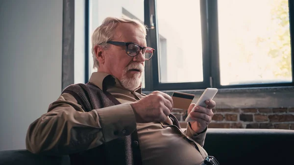 Pensioner in eyeglasses using smartphone and holding credit card at home — Stock Photo