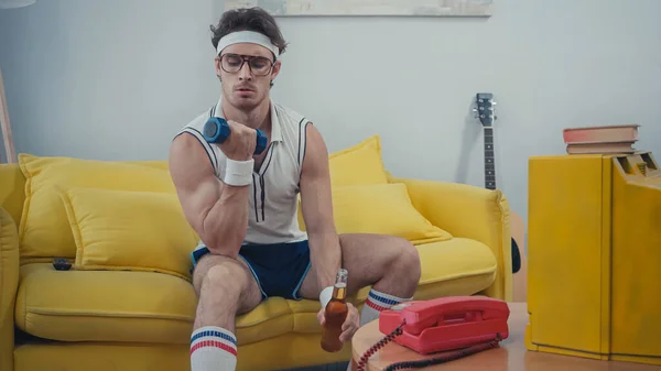 Sportsman holding glass bottle of beer and lifting dumbbell while sitting on sofa — Stock Photo