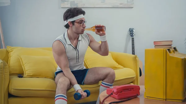 Sportsman drinking beer and lifting dumbbell while sitting on sofa — Stock Photo