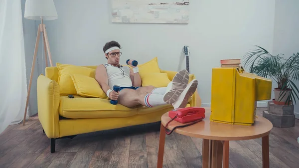 Man in eyeglasses training with dumbbells while sitting on couch and watching tv in living room — Stock Photo