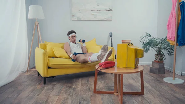 Hombre con gafas haciendo ejercicio con pesas mientras está sentado en el sofá y viendo la televisión en la sala de estar - foto de stock
