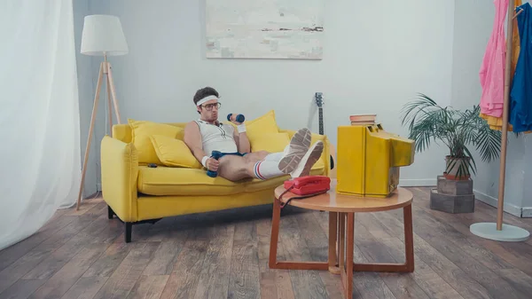Man in eyeglasses exercising with dumbbells while sitting on couch and watching tv in living room — Stock Photo