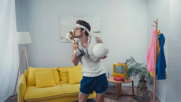 Esportista beijando copo troféu e bola de esportes enquanto está em casa — Fotografia de Stock