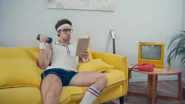 Sportsman lifting dumbbell and pouting lips while reading book on sofa in living room — Stock Photo