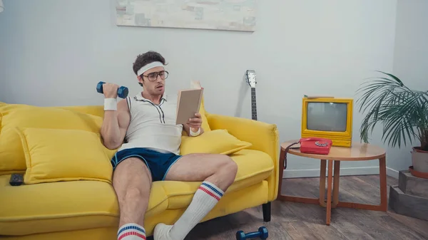 Sportsman lifting dumbbell while reading book on sofa in living room — Stock Photo