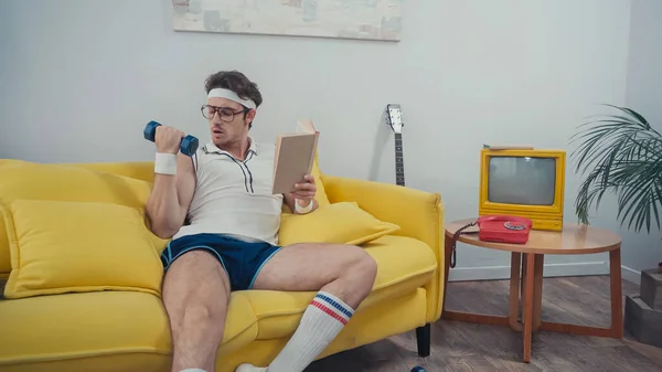 Sportsman lifting dumbbell while holding book in living room — Stock Photo