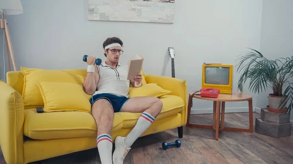 Sportsman lifting dumbbell and reading book in living room — Stock Photo