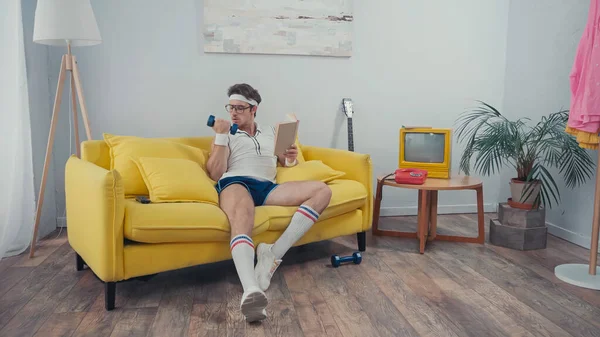 Sportsman exercising with dumbbell while holding book in living room — Stock Photo