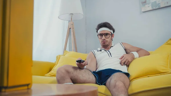 Man in retro sportswear sitting on couch and switching channels — Stock Photo