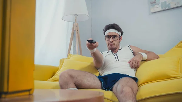Man in retro sportswear sitting on yellow sofa and switching channels — Stock Photo