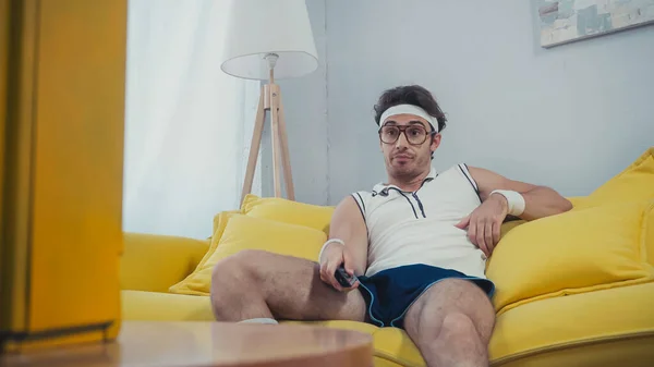 Недовольный мужчина в ретро спортивной одежде сидит на диване и смотрит телевизор — стоковое фото