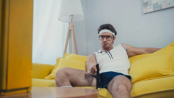 Молодий нудний чоловік в окулярах і спортивному одязі сидить на дивані і комутаційних каналах — стокове фото