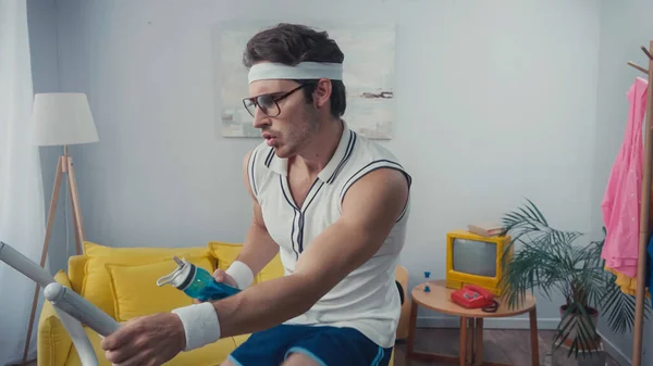 Sportsman working out on exercise bike and holding sports bottle in living room, retro sport concept — Stock Photo