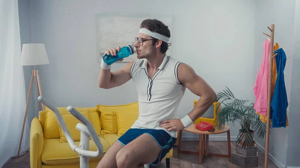 Deportista haciendo ejercicio en bicicleta estática y agua potable en la sala de estar, concepto de deporte retro - foto de stock