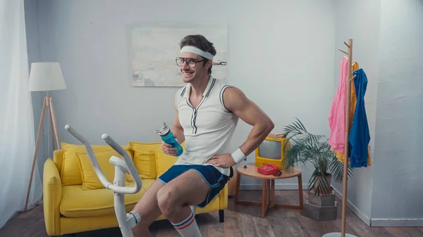 Entrenamiento de deportista alegre en bicicleta estática y celebración de botella deportiva en la sala de estar, concepto de deporte retro - foto de stock