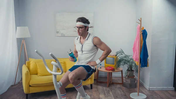 Втомлений спортсмен тренується на тренажерах велосипеді і тримає спортивну пляшку у вітальні, концепція ретро спорту — стокове фото
