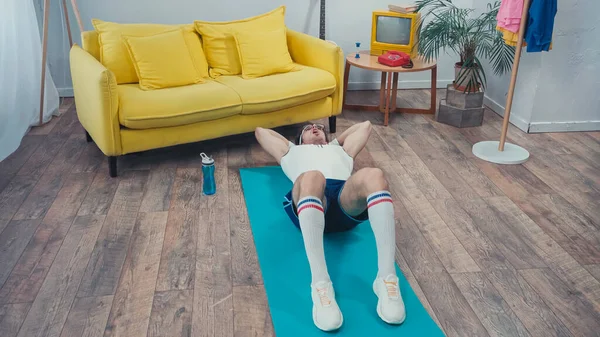 Deportista cansado en gafas haciendo ejercicios abdominales en la alfombra de fitness en la sala de estar - foto de stock