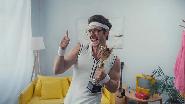 Sportsman in eyeglasses showing thumb up and holding golden trophy at home — Stock Photo