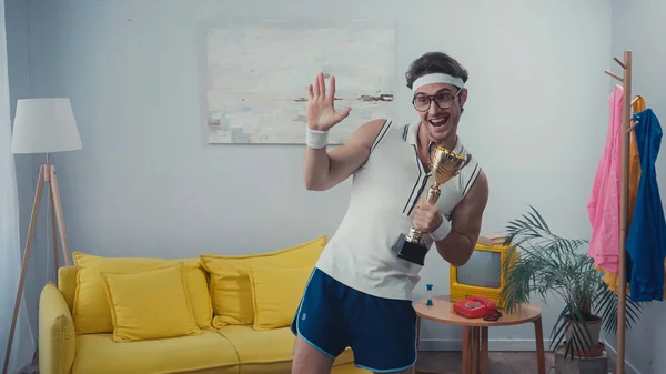 Happy sportsman in eyeglasses waving hand and holding golden trophy at home — Stock Photo