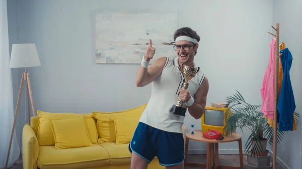 Deportista alegre en gafas con trofeo de oro y el pulgar hacia arriba en casa - foto de stock