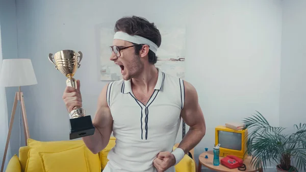 Excited sportsman in eyeglasses screaming while holding golden trophy at home — Stock Photo