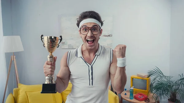 Amazed winner in sportswear and eyeglasses holding golden trophy at home — Stock Photo