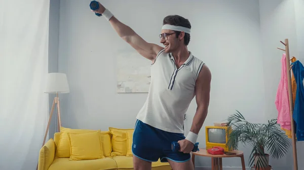 Desportista alegre levantando halteres e dançando na sala de estar, conceito de esporte retro — Fotografia de Stock