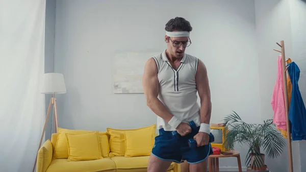 Desportista em óculos exercitando com halteres na sala de estar, conceito de esporte retro — Fotografia de Stock