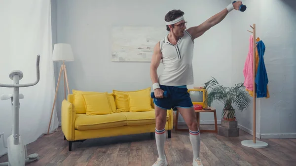 Esportista engraçado em óculos exercitando com halteres na sala de estar, conceito de esporte retro — Fotografia de Stock