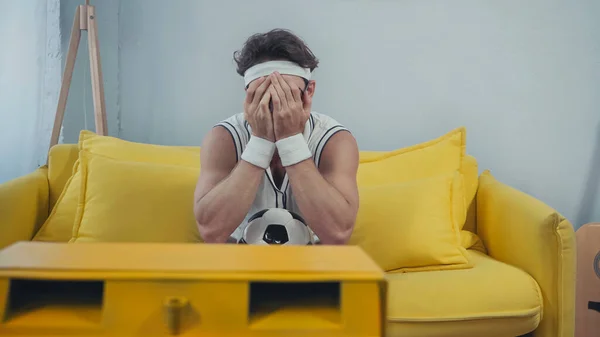 Незадоволений футбольний вентилятор у спортивному одязі, що закриває обличчя під час перегляду телевізора на дивані — стокове фото